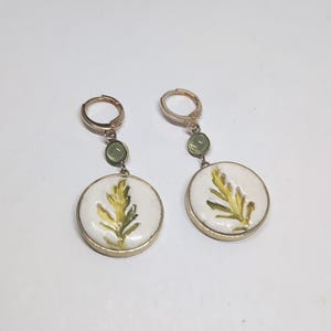 May include: A pair of gold-toned hoop earrings featuring a small, round, light green stone accent above a larger, circular white pendant. Each pendant displays a painted yellow and green leaf design encased in a thin gold-toned metal frame.