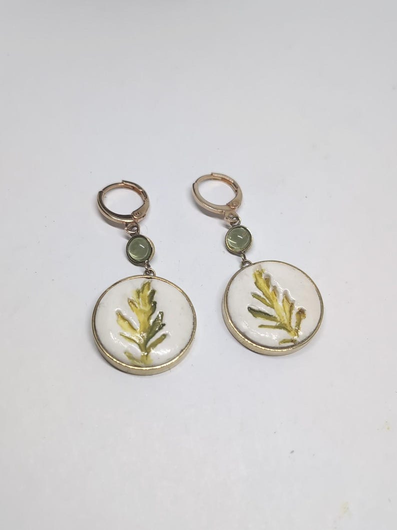 May include: A pair of gold-toned hoop earrings featuring a small, round, light green stone accent above a larger, circular white pendant. Each pendant displays a painted yellow and green leaf design encased in a thin gold-toned metal frame.