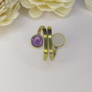 May include: A bypass ring crafted from a brushed brass-toned metal, featuring two open ends. One end is set with a circular, deep purple stone, while the other end holds a circular, smooth, opaque white stone, creating a contrasting dual-stone design.
