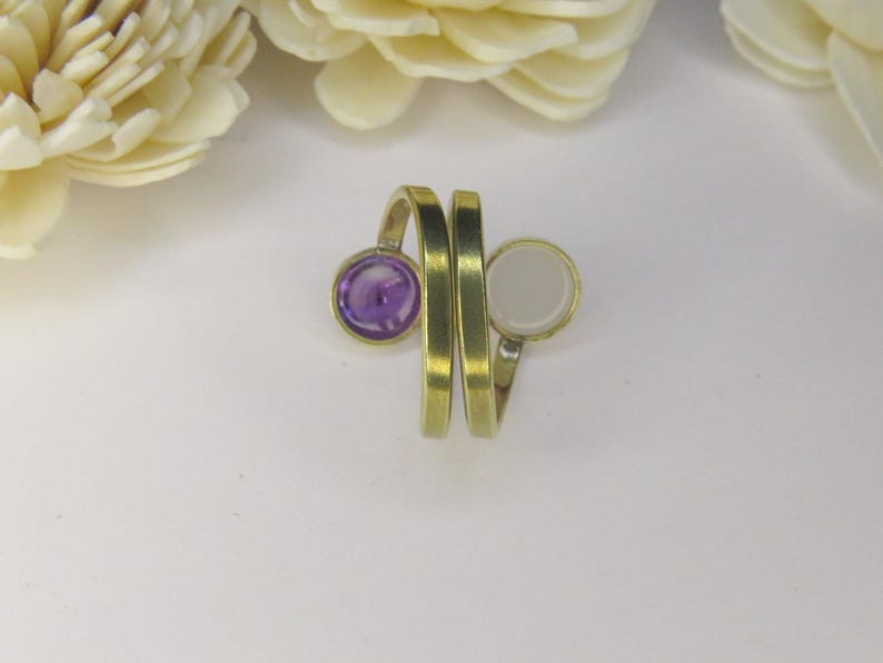 May include: A bypass ring crafted from a brushed brass-toned metal, featuring two open ends. One end is set with a circular, deep purple stone, while the other end holds a circular, smooth, opaque white stone, creating a contrasting dual-stone design.