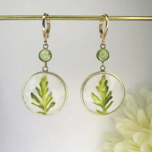 May include: A pair of gold-toned dangle earrings featuring a circular white pendant with a green leaf illustration. Each earring includes a small round green accent stone above the pendant and a gold-toned lever-back closure for secure attachment.