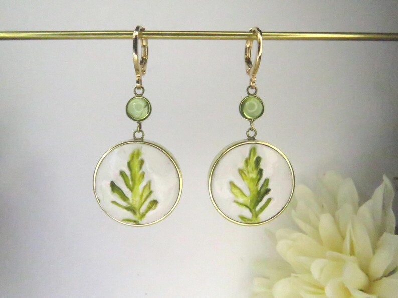 May include: A pair of gold-toned dangle earrings featuring a circular white pendant with a green leaf illustration. Each earring includes a small round green accent stone above the pendant and a gold-toned lever-back closure for secure attachment.