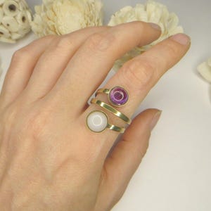 May include: A gold-toned bypass ring featuring two circular settings. One setting contains a translucent purple stone, while the other holds an opaque white stone. The band has an open, overlapping design that wraps around the finger.