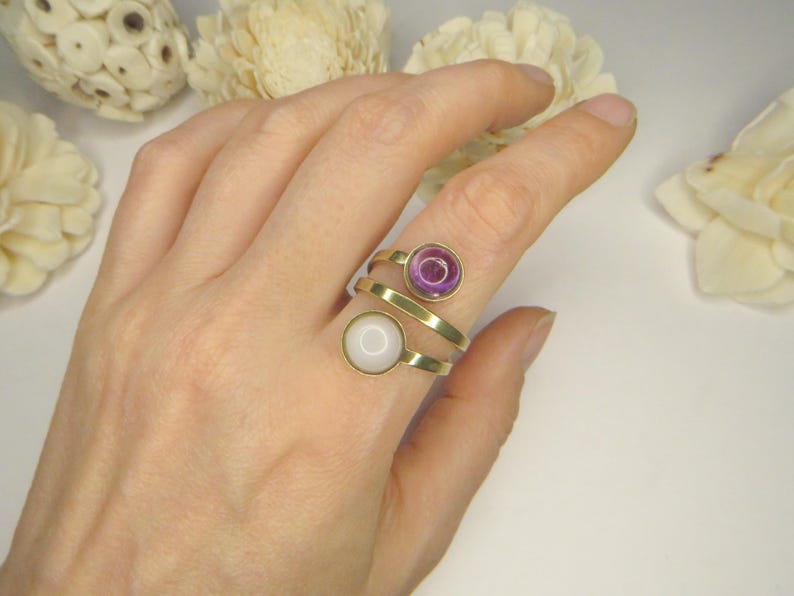 May include: A gold-toned bypass ring featuring two circular settings. One setting contains a translucent purple stone, while the other holds an opaque white stone. The band has an open, overlapping design that wraps around the finger.