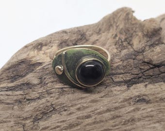 Op de afbeelding: Een ring met een ronde zwarte steen in een metalen band met een groene patina afwerking. Het ontwerp bevat een klein goudkleurig rond accent aan de zijkant, verbonden door een dunne gebogen lijn met de zetting van de centrale steen op de band.