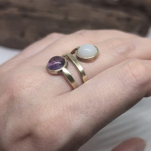May include: A gold-toned bypass ring featuring two circular settings. One setting contains a smooth, translucent purple cabochon stone, while the other setting holds a smooth, opaque white cabochon stone. The band is made of a simple, polished metal.