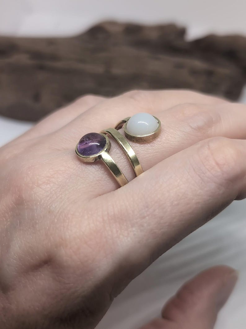 May include: A gold-toned bypass ring featuring two circular settings. One setting contains a smooth, translucent purple cabochon stone, while the other setting holds a smooth, opaque white cabochon stone. The band is made of a simple, polished metal.