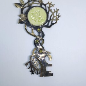 May include: A decorative metal key-shaped brooch featuring a circular pale green cabochon with a frog relief. The metalwork has a dark oxidized finish with polished gold-tone accents, leaf motifs, and three small embedded green stones.