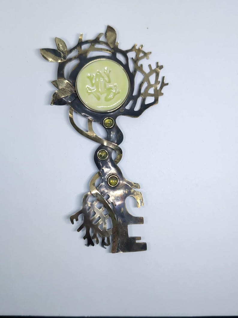 May include: A decorative metal key-shaped brooch featuring a circular pale green cabochon with a frog relief. The metalwork has a dark oxidized finish with polished gold-tone accents, leaf motifs, and three small embedded green stones.