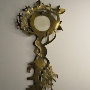 May include: A brass wall hanging shaped like a key with an intricate, organic design. The top features a circular opening with a white insert, surrounded by branch-like cutouts. The stem is twisted, and the bottom resembles a root system.