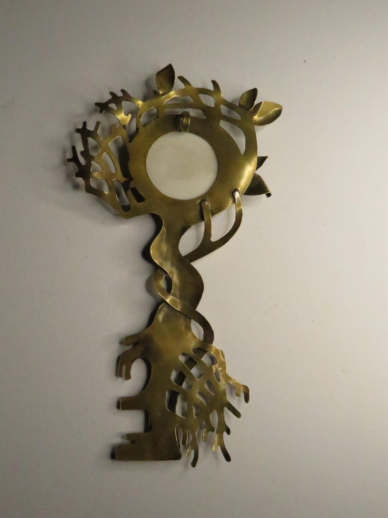 May include: A brass wall hanging shaped like a key with an intricate, organic design. The top features a circular opening with a white insert, surrounded by branch-like cutouts. The stem is twisted, and the bottom resembles a root system.