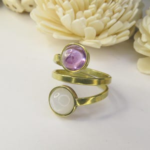 May include: A gold-toned bypass ring featuring two circular cabochon stones. The top stone is a translucent light purple, while the bottom stone is an opaque white. The metal band has a smooth, polished finish and wraps around the finger.
