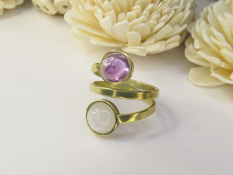 May include: A gold-toned bypass ring featuring two circular cabochon stones. The top stone is a translucent light purple, while the bottom stone is an opaque white. The metal band has a smooth, polished finish and wraps around the finger.
