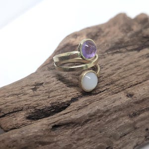 May include: A bypass ring crafted from gold-toned metal featuring two round bezel-set stones. One stone is a translucent purple amethyst, and the other is an opaque white moonstone. The band wraps around the finger in a spiral design.