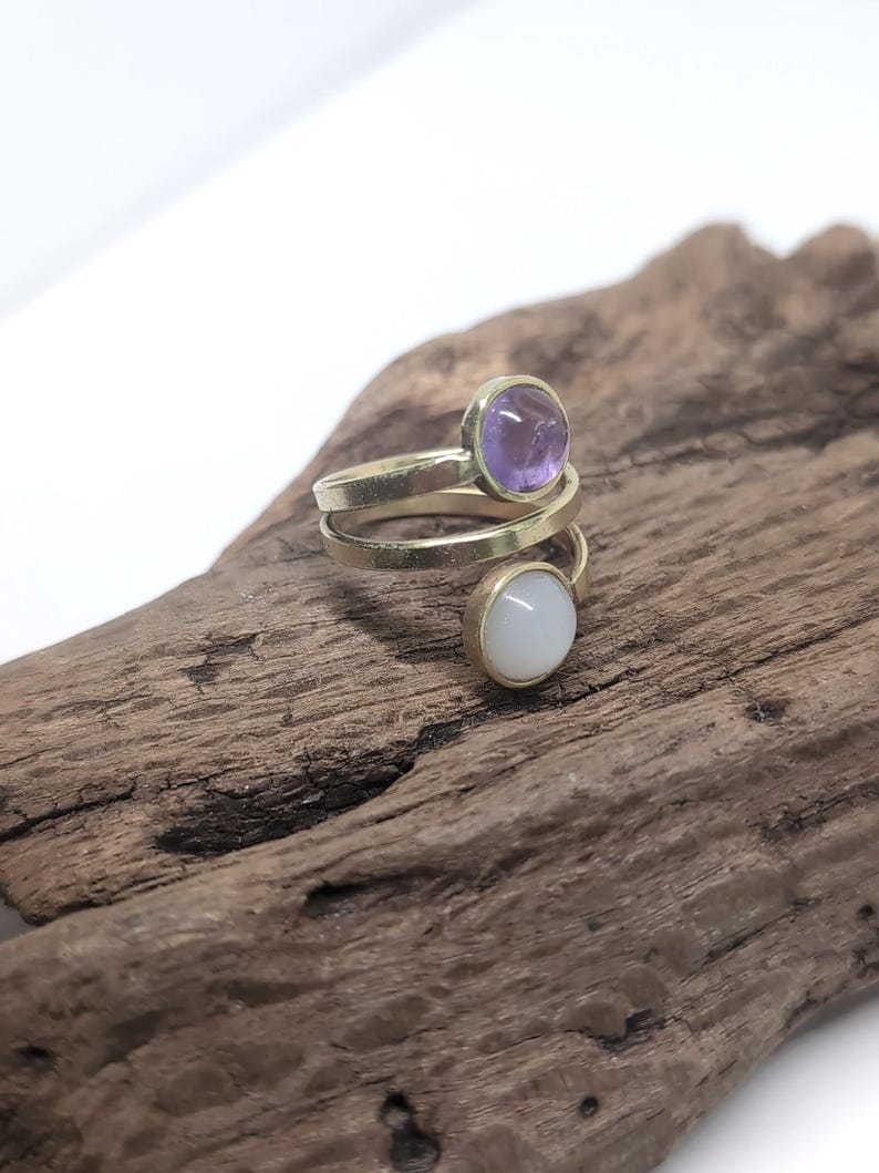 May include: A bypass ring crafted from gold-toned metal featuring two round bezel-set stones. One stone is a translucent purple amethyst, and the other is an opaque white moonstone. The band wraps around the finger in a spiral design.