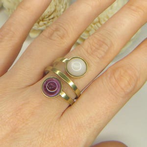 May include: A gold-toned bypass ring featuring two circular settings. One setting contains a smooth, opaque white stone, while the other holds a translucent, deep purple circular stone. The band is made of a polished, flat gold-colored metal.
