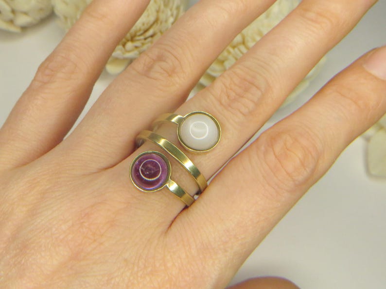 May include: A gold-toned bypass ring featuring two circular settings. One setting contains a smooth, opaque white stone, while the other holds a translucent, deep purple circular stone. The band is made of a polished, flat gold-colored metal.