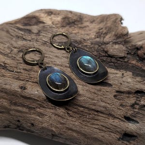 May include: A pair of drop earrings featuring oval labradorite gemstones with blue iridescence, set in brass bezels on dark, teardrop-shaped metal backings. The earrings are attached to circular brass lever-back closures for secure fastening.