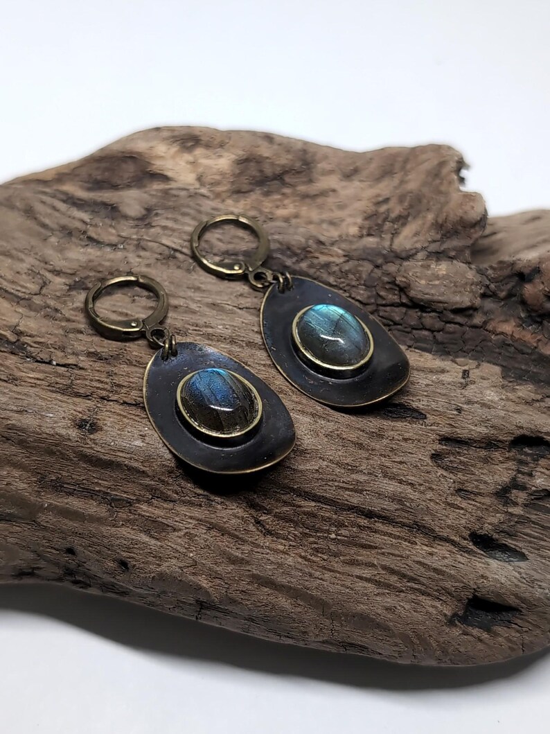 May include: A pair of drop earrings featuring oval labradorite gemstones with blue iridescence, set in brass bezels on dark, teardrop-shaped metal backings. The earrings are attached to circular brass lever-back closures for secure fastening.
