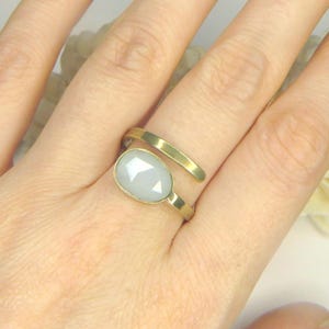 May include: A gold-toned bypass ring featuring a smooth, polished metal band that wraps around the finger. The ring is accented with a single, oval-shaped, faceted light gray stone set in a simple gold-toned bezel setting on one end.