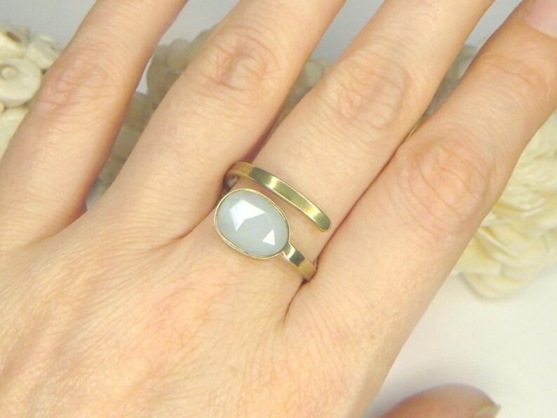 May include: A gold-toned bypass ring featuring a smooth, polished metal band that wraps around the finger. The ring is accented with a single, oval-shaped, faceted light gray stone set in a simple gold-toned bezel setting on one end.