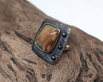 May include: A ring featuring a rectangular, multi-colored stone set in a gold-toned bezel, mounted on a dark, oxidized metal plate with a textured, swirling pattern and three small, raised metal spheres on the right side.