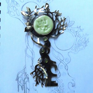 May include: A metal brooch featuring a dark, oxidized finish with a tree-like structure. It includes a round, pale green ceramic cabochon at the top and three small, round, green faceted gemstones set vertically along the central stem.
