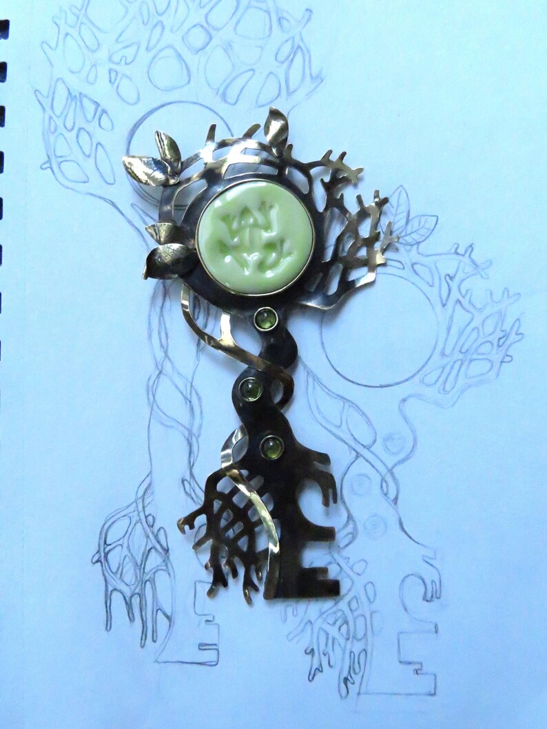 May include: A metal brooch featuring a dark, oxidized finish with a tree-like structure. It includes a round, pale green ceramic cabochon at the top and three small, round, green faceted gemstones set vertically along the central stem.
