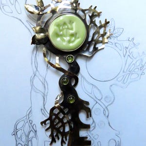 May include: A metal brooch featuring a dark, oxidized finish with a tree-like structure. It includes a round, light green cabochon with a relief frog design, three small green faceted stones, and polished metal leaves at the top.