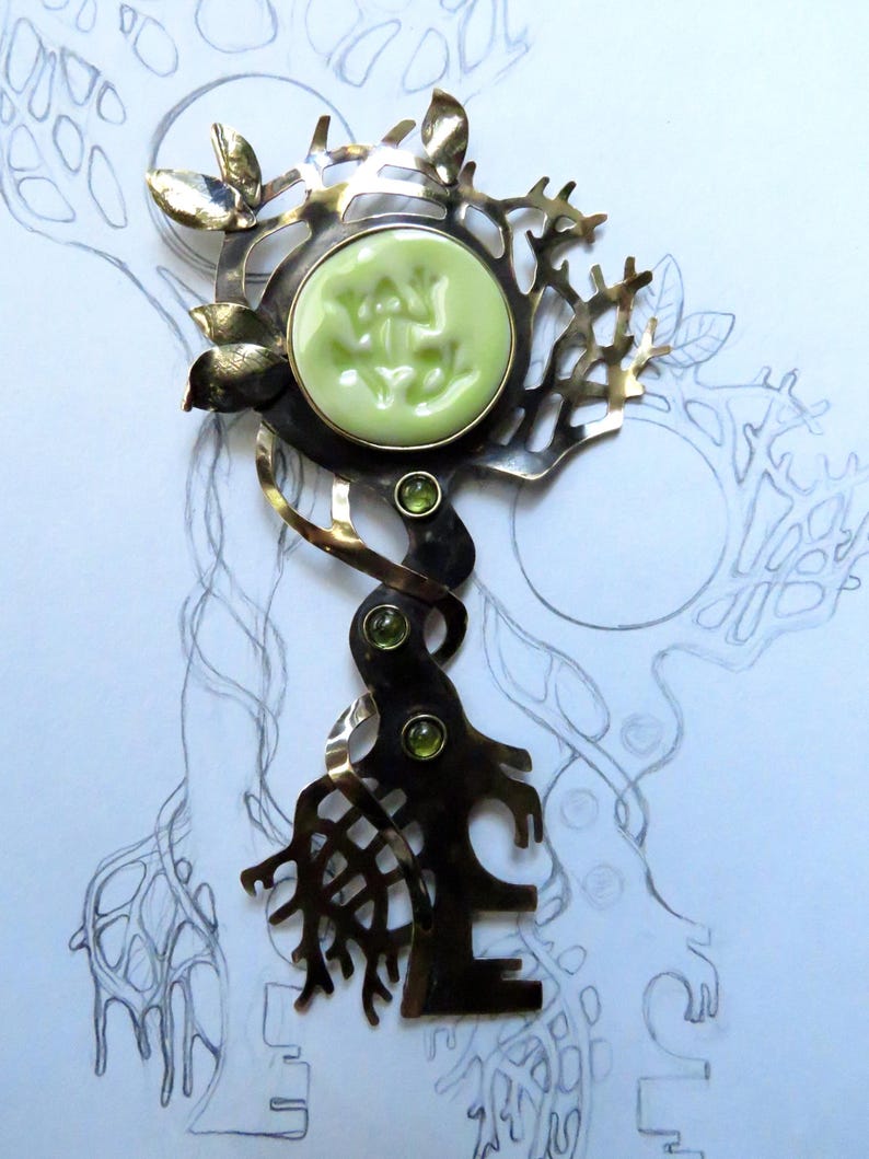 May include: A metal brooch featuring a dark, oxidized finish with a tree-like structure. It includes a round, light green cabochon with a relief frog design, three small green faceted stones, and polished metal leaves at the top.