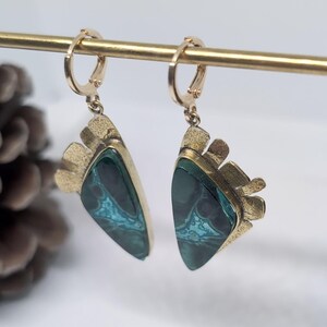 May include: A pair of dangle earrings featuring triangular-shaped stones with dark green and teal marbled patterns. The stones are set in textured gold-toned metal frames with a fan-like detail at the top, suspended from small gold-toned hoop clasps.