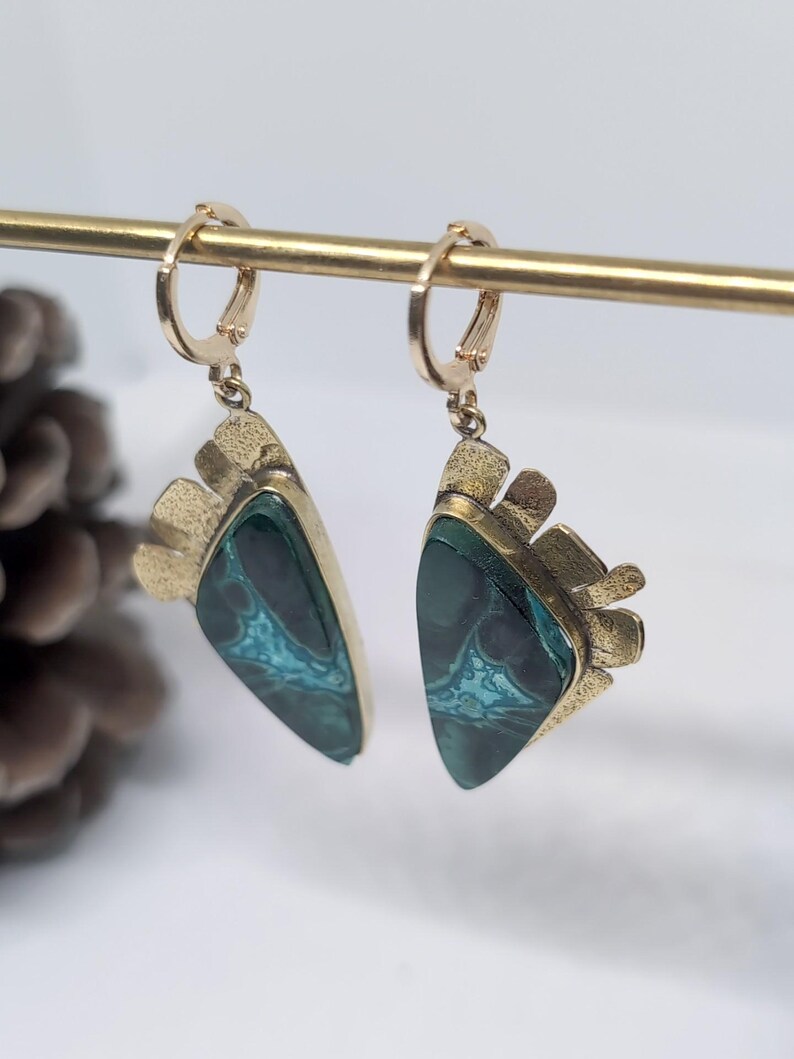 May include: A pair of dangle earrings featuring triangular-shaped stones with dark green and teal marbled patterns. The stones are set in textured gold-toned metal frames with a fan-like detail at the top, suspended from small gold-toned hoop clasps.