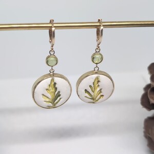 May include: A pair of gold-toned drop earrings featuring a circular white pendant with a green leaf illustration. Each earring includes a small, round, light green gemstone accent positioned above the circular pendant, suspended from a gold hoop.