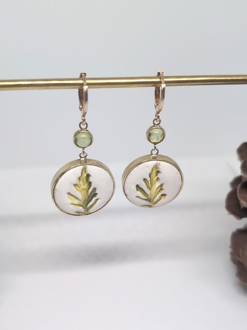 May include: A pair of gold-toned drop earrings featuring a circular white pendant with a green leaf illustration. Each earring includes a small, round, light green gemstone accent positioned above the circular pendant, suspended from a gold hoop.
