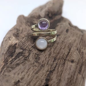 May include: A bypass ring featuring a gold-toned metal band that wraps around the finger. The ring is set with two circular cabochon stones: one translucent light purple stone at the top and one opaque white stone at the bottom.