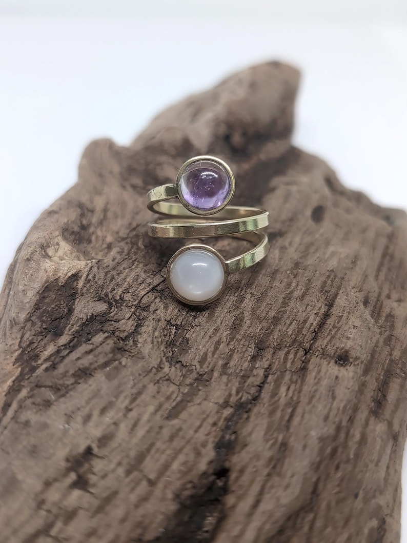May include: A bypass ring featuring a gold-toned metal band that wraps around the finger. The ring is set with two circular cabochon stones: one translucent light purple stone at the top and one opaque white stone at the bottom.