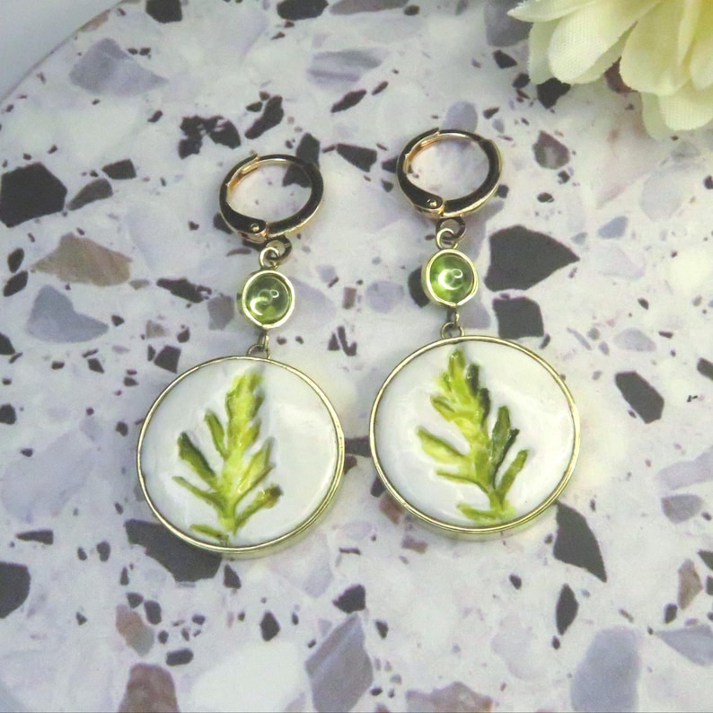 May include: A pair of gold-toned dangle earrings featuring circular white pendants with a green leaf illustration. Each earring has a small green accent stone set above the pendant and a gold hoop closure at the top for secure attachment.