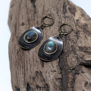 May include: A pair of drop earrings featuring oval labradorite gemstones set in brass bezels. The stones are mounted on dark, oxidized metal discs with a smooth, rounded shape. Each earring is attached to a brass lever-back closure mechanism.