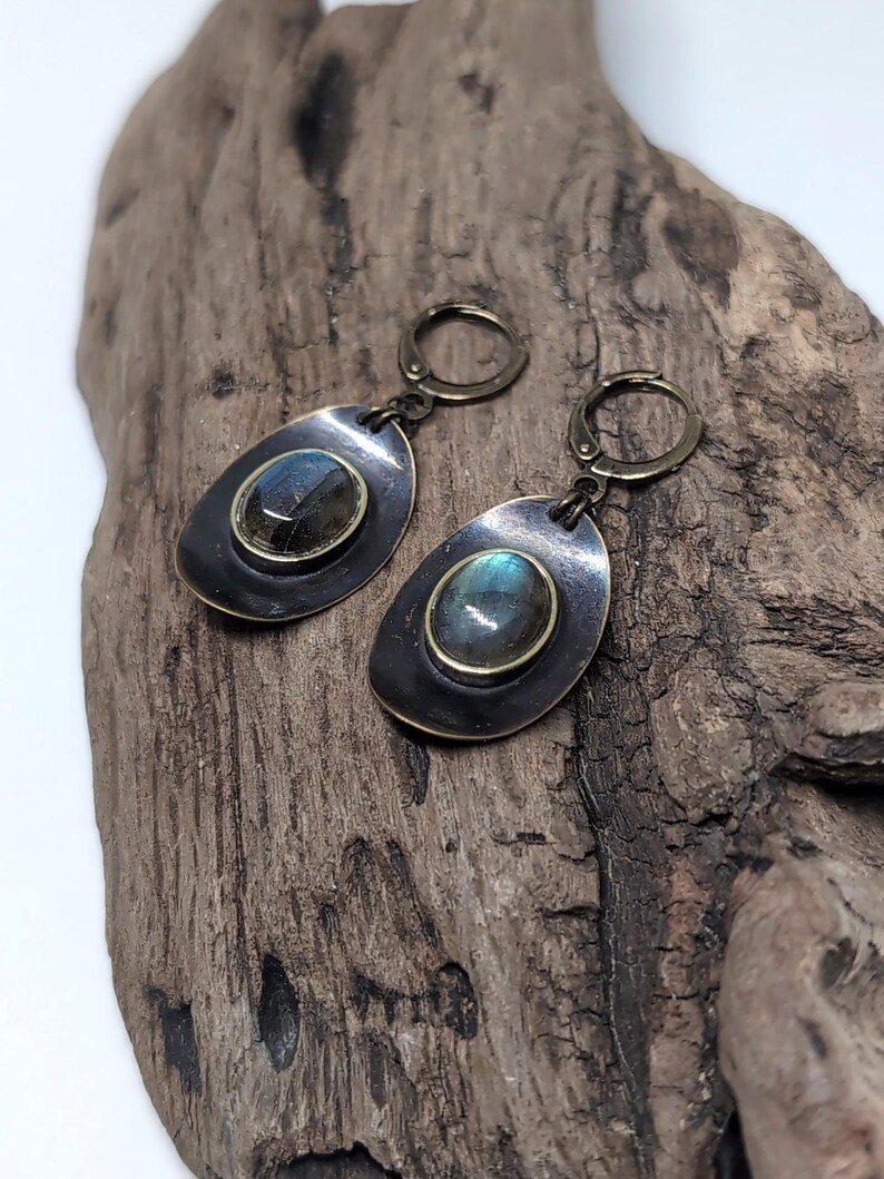 May include: A pair of drop earrings featuring oval labradorite gemstones set in brass bezels. The stones are mounted on dark, oxidized metal discs with a smooth, rounded shape. Each earring is attached to a brass lever-back closure mechanism.