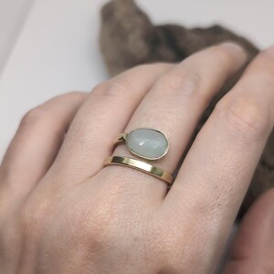 May include: A gold-toned bypass ring featuring a smooth, polished band and a second band topped with a faceted, oval-shaped pale green stone. The ring has an open design where the two ends do not meet, showcasing a clean and simple metal finish.
