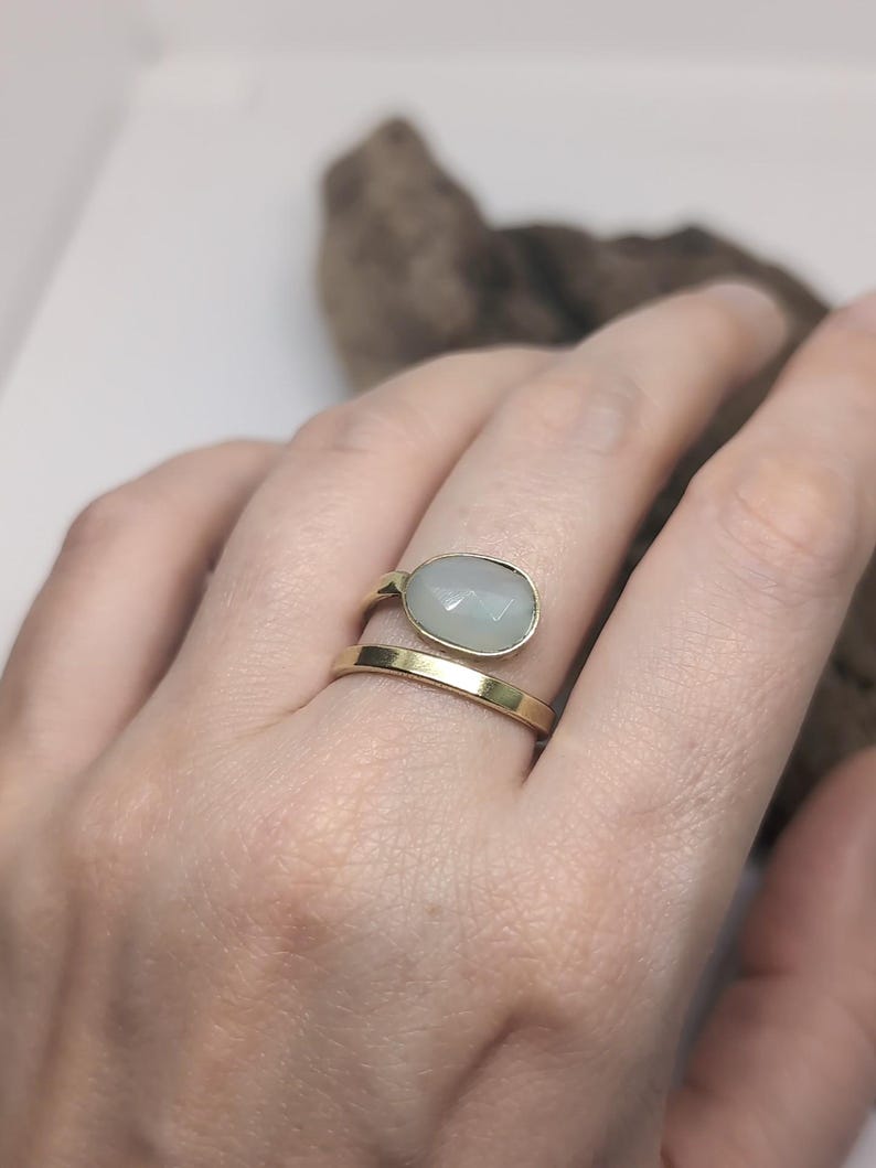 May include: A gold-toned bypass ring featuring a smooth, polished band and a second band topped with a faceted, oval-shaped pale green stone. The ring has an open design where the two ends do not meet, showcasing a clean and simple metal finish.