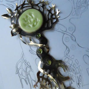 May include: A metal key-shaped brooch featuring a dark, oxidized finish with branch-like cutouts and small leaves. The head of the key holds a round, pale green cabochon with embossed text, while the stem is accented with three small green stones.