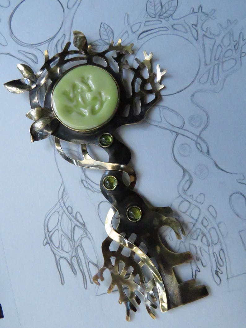 May include: A metal key-shaped brooch featuring a dark, oxidized finish with branch-like cutouts and small leaves. The head of the key holds a round, pale green cabochon with embossed text, while the stem is accented with three small green stones.