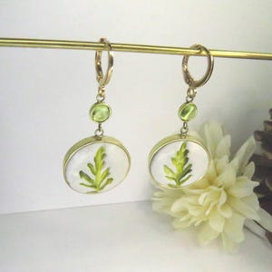 May include: A pair of gold-toned dangle earrings featuring a circular pendant with a white background and a green fern leaf illustration. Each earring includes a small round green accent stone above the pendant and a gold-toned hoop closure.