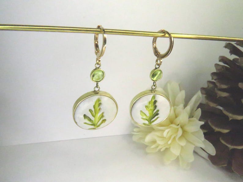 May include: A pair of gold-toned dangle earrings featuring a circular pendant with a white background and a green fern leaf illustration. Each earring includes a small round green accent stone above the pendant and a gold-toned hoop closure.
