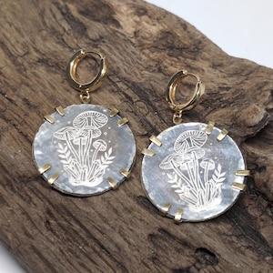 May include: A pair of circular earrings featuring a grey, iridescent shell-like inlay with a white etched design of mushrooms and crystals. Each disc is secured in a gold-toned metal setting with four prongs and hangs from a gold-toned hoop.