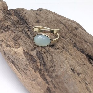 May include: A gold-toned bypass ring featuring a single oval-shaped, faceted, pale blue-green stone set in a smooth metal bezel. The band is open and wraps around the finger, showcasing a polished metallic finish and a soft, translucent gemstone.