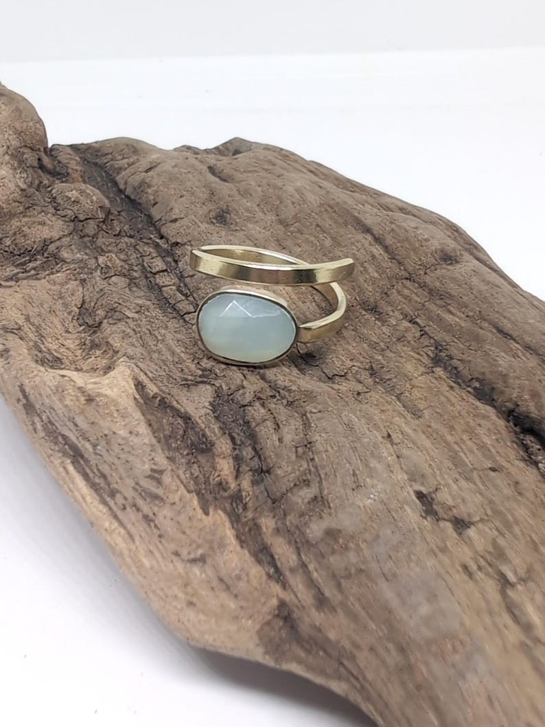 May include: A gold-toned bypass ring featuring a single oval-shaped, faceted, pale blue-green stone set in a smooth metal bezel. The band is open and wraps around the finger, showcasing a polished metallic finish and a soft, translucent gemstone.