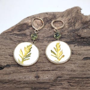 May include: A pair of gold-toned dangle earrings featuring circular white pendants with a painted yellow and green fern leaf design. Each earring includes a small, round, translucent green bead accent and a secure lever-back closure mechanism.