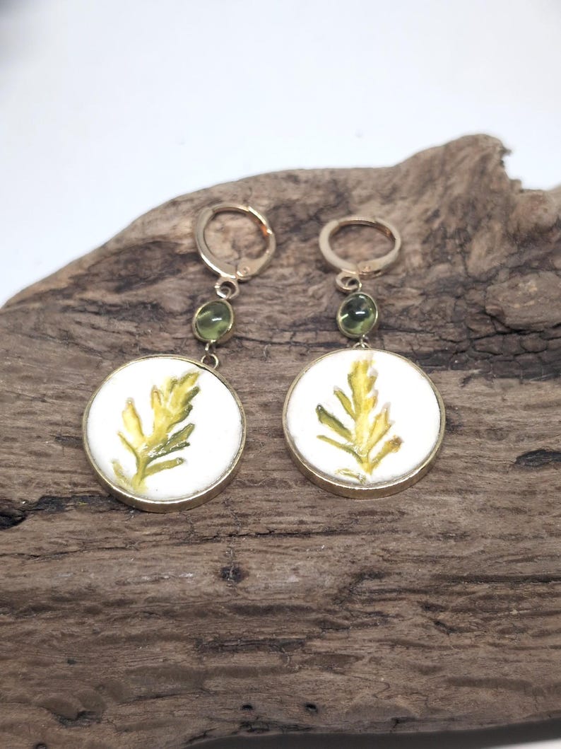 May include: A pair of gold-toned dangle earrings featuring circular white pendants with a painted yellow and green fern leaf design. Each earring includes a small, round, translucent green bead accent and a secure lever-back closure mechanism.